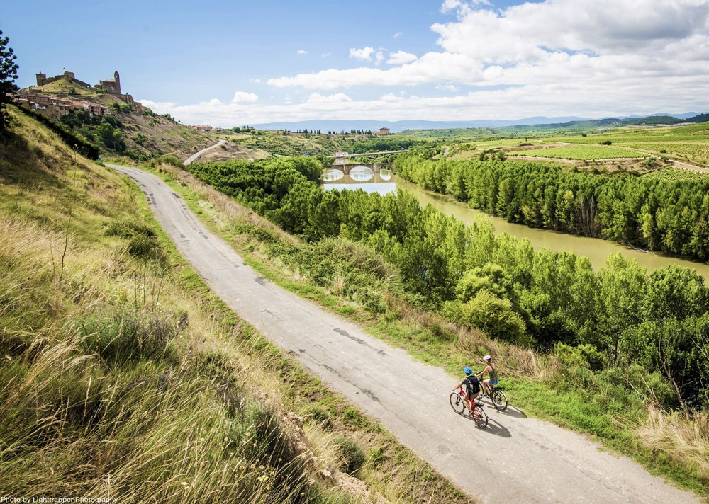 Two cyclists ascend a quiet country lane towards a hill fort in Rioja