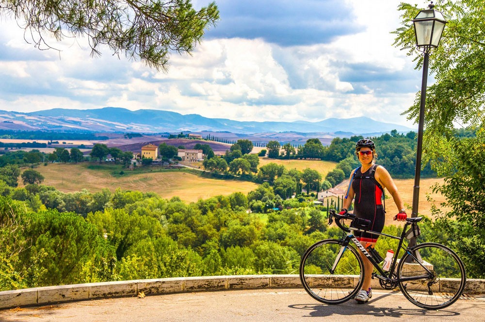 A woman stands with her bike against a picturesque backdrop of Tuscany's hills